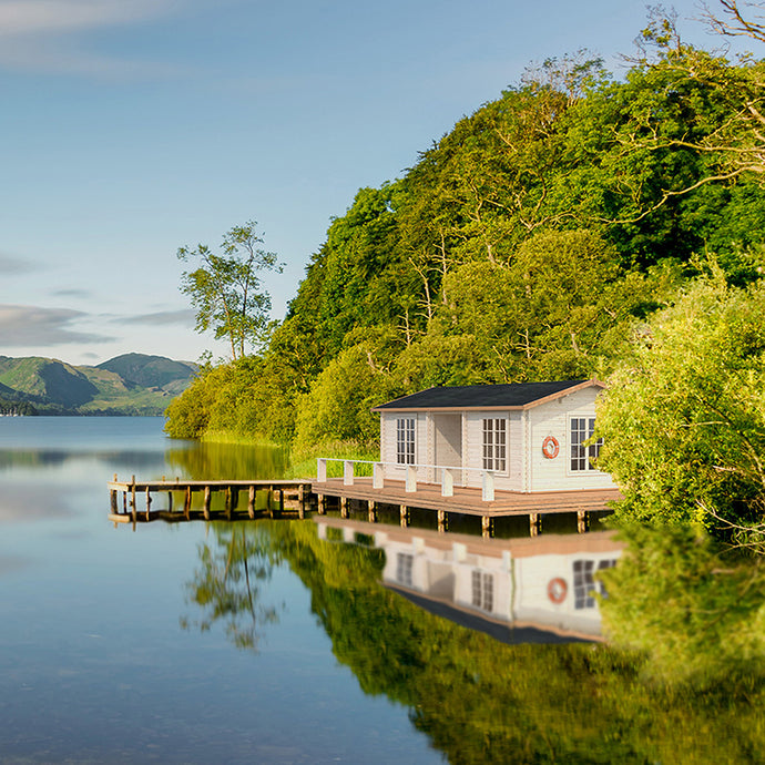 A serene lakeside setting featuring the Cabin New Zealand 343 Sq.ft. The cabin is positioned on a wooden deck with a view of the water, surrounded by lush greenery, creating the perfect retreat by WholeWoodCabins